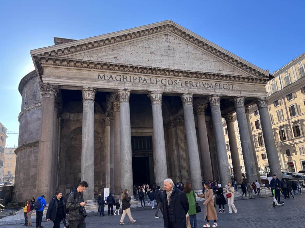 Front view of the Pantheon in Rome, showcasing its ancient Roman architecture with Corinthian columns and a pediment inscribed with Latin text, with visitors milling around the entrance.