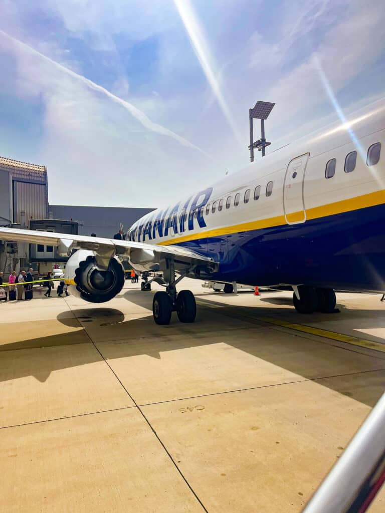 This image captures a Ryanair airplane on the tarmac, viewed from a side angle from boarding at the back. The plane's livery, featuring dark blue and yellow, is vibrant under the bright, clear sky. Ground personnel can be seen near the aircraft. Sunlight casts strong reflections and shadows, adding contrast to the scene.




