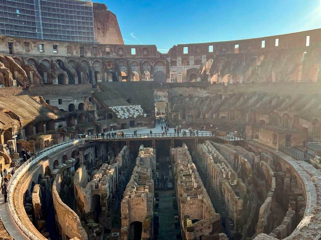 Late afternoon sun highlights the depth and detail of the Colosseum's internal ruins, with the maze-like underground visibly intricate, and visitors exploring the historic site.