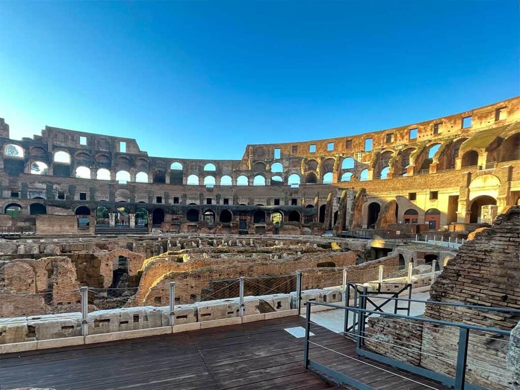 Morning light bathes the Colosseum's interior ruins, highlighting the complex underground chambers and the grandeur of the arches against a clear blue sky.
