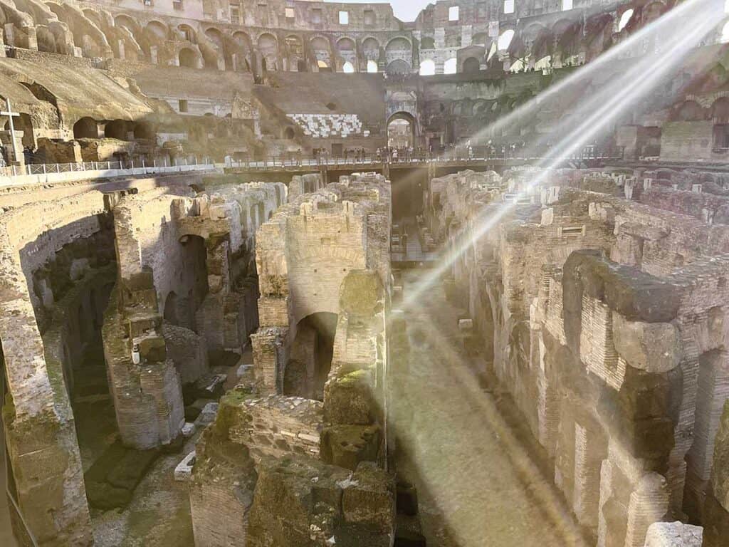 Shafts of sunlight pierce through the Colosseum's upper levels, casting a mystical glow over the ancient underground ruins and the impressive array of arches above
