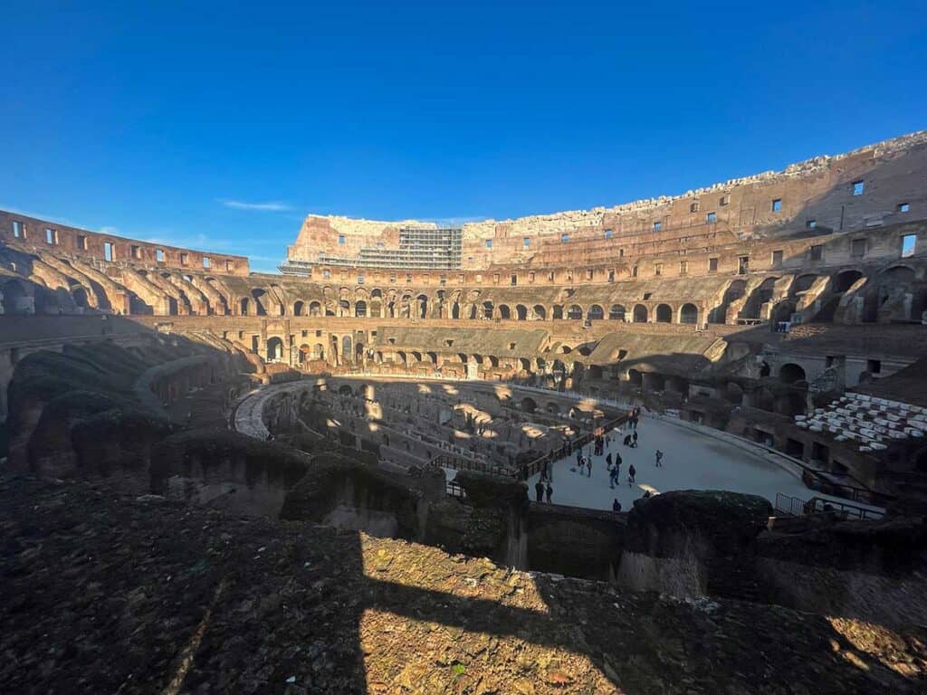 Inside the Colosseum, capturing the vast interior with its historic layered walls and arches, bathed in sunlight, with visitors scattered on the ancient arena floor.