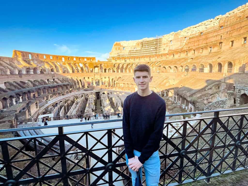 A young person with a content smile, wearing a dark sweater, standing before the grand interior backdrop of the Colosseum during the day during a Colosseum tour, with the ancient arena's ruins visible behind a protective railing.