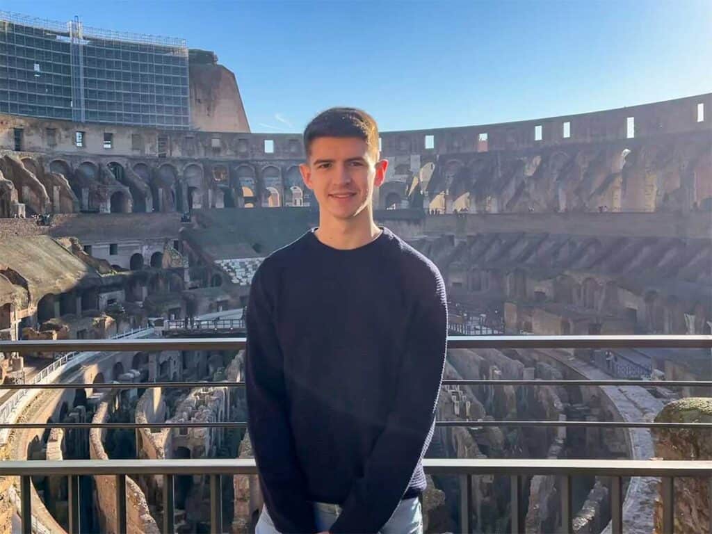 A confident young person in a dark sweater stands with the Colosseum's impressive ancient arena and rows of arches in the background, under a sunlit sky