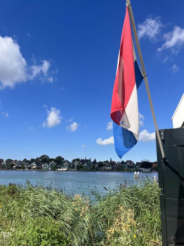 A picture of a Dutch flag on the side of a building in front of some reeds and a body of water with traditional Dutch houses on the other side. 