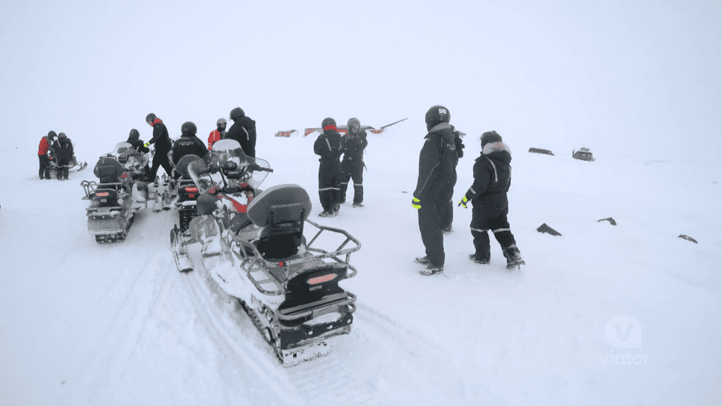 A group of snowmobiles with some tour participants standing around. Taking a snowmobile tour in Iceland is an incredible experience.