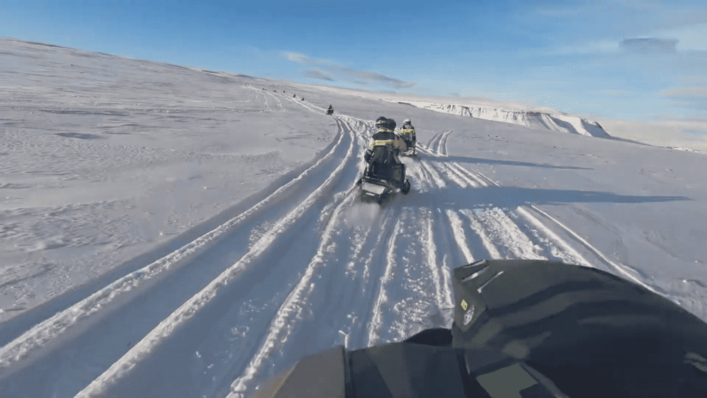 A picture taken from a snowmobile where you can see other snowmobiles ahead as they drive through the snow during a snowmobile tour in Iceland.