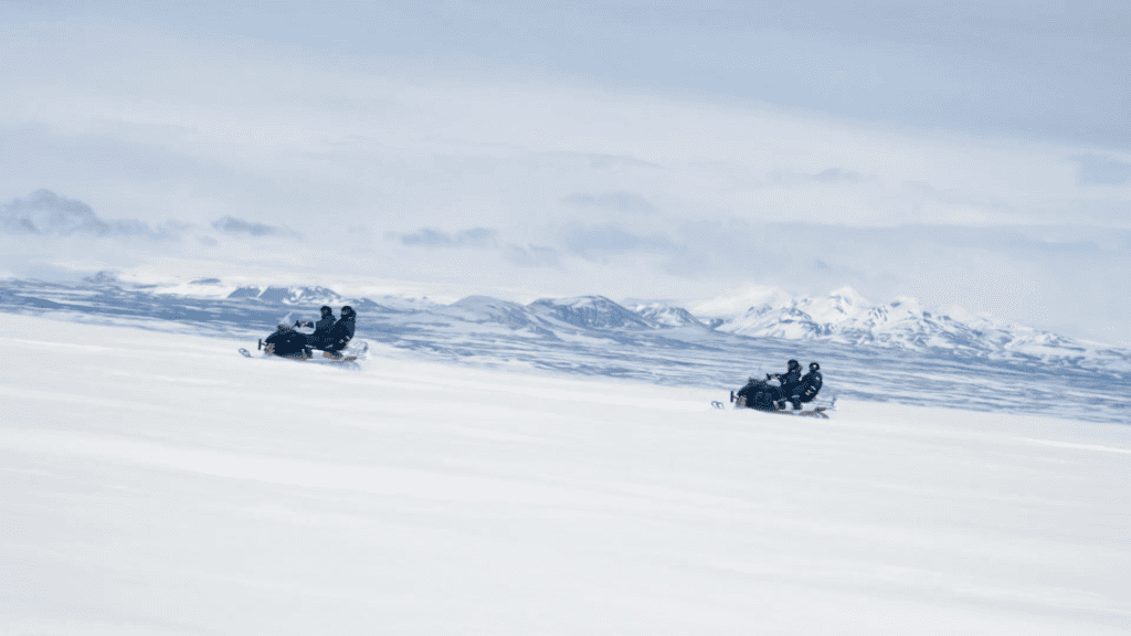 Two snowmobiles driving with two people on each snowmobile past some snowy mountains in the distance.