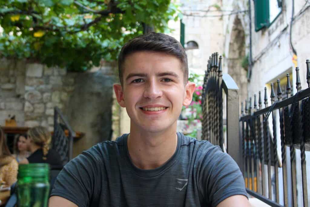 Smiling young man with short dark hair sitting at an outdoor restaurant with green foliage and stone walls in the background