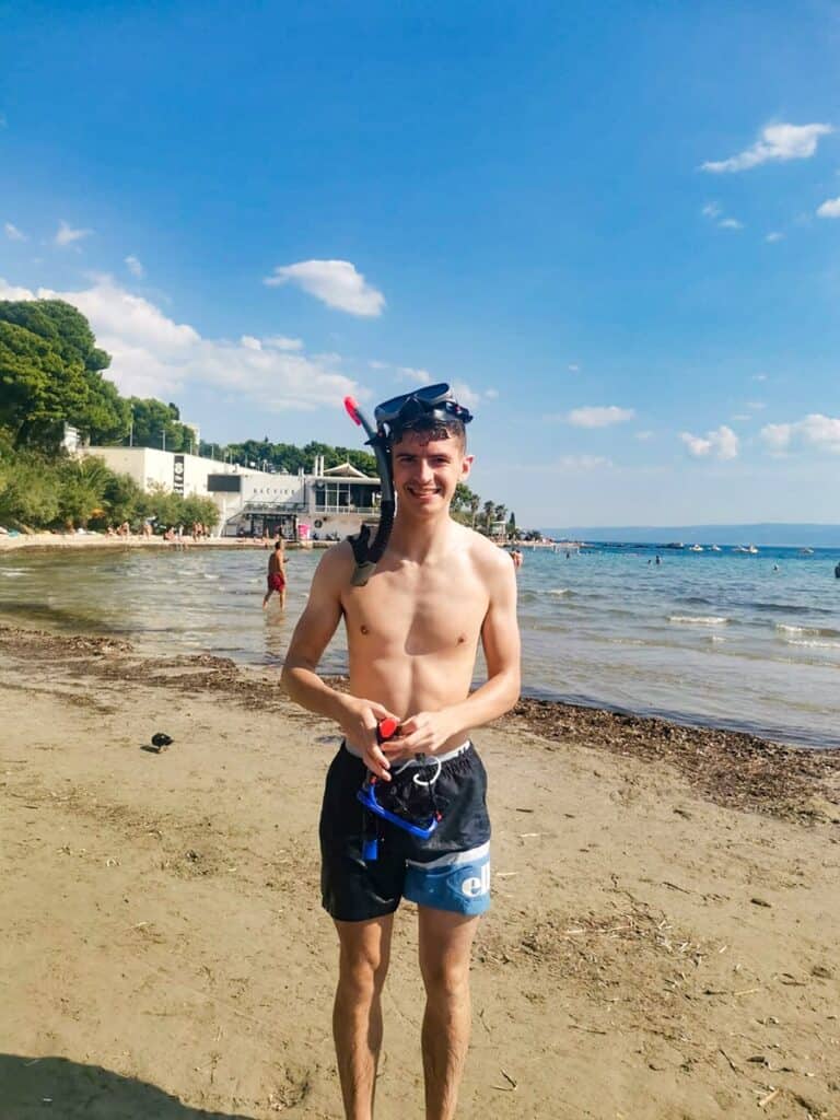 Young man ready for snorkeling wearing a snorkel and fins, with the beach and holidaymakers in the background.