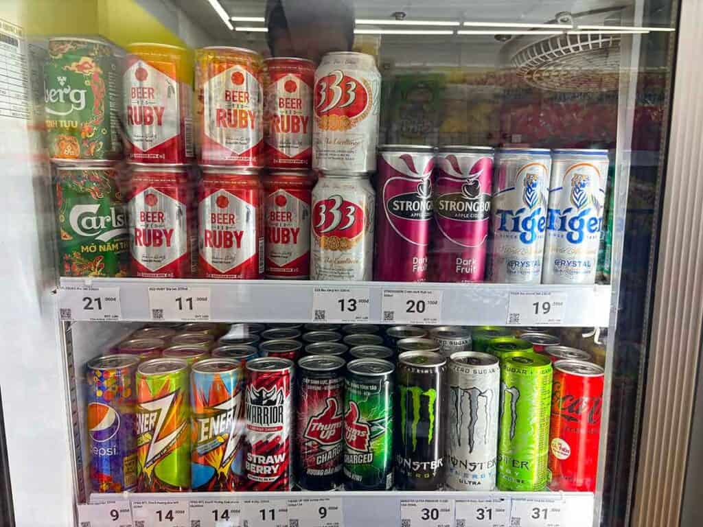 An assortment of canned and bottled beverages neatly arranged in a refrigerated shelf, including beers and energy drinks, with price tags in Thai Baht.