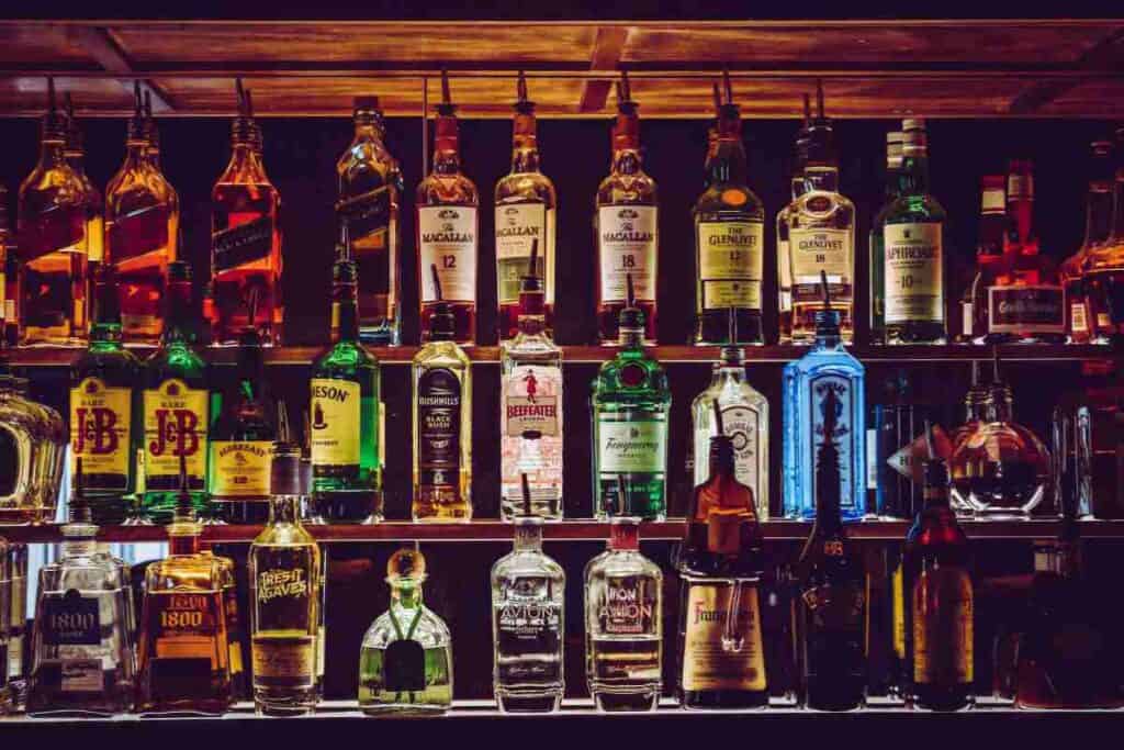 A dimly lit shelf stocked with a variety of spirits and liquors in a cozy bar setting, showcasing bottles of different shapes and labels.
