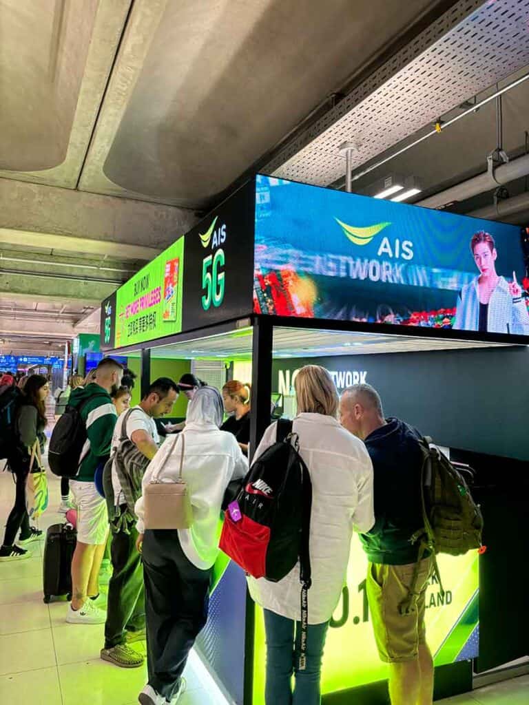 Travelers queue at a brightly lit airport SIM card stall with prominent 'AIS 5G' branding above the counter