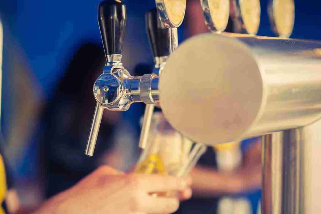 A close-up of a beer tap with a glass being filled with golden ale, set against a blurred bar backdrop. The drinking age in Thailand is 20.