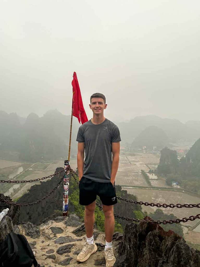 A man stands proudly with a red flag at the peak of Mua Cave overlooking the misty landscape of Ninh Binh Province, Vietnam.
