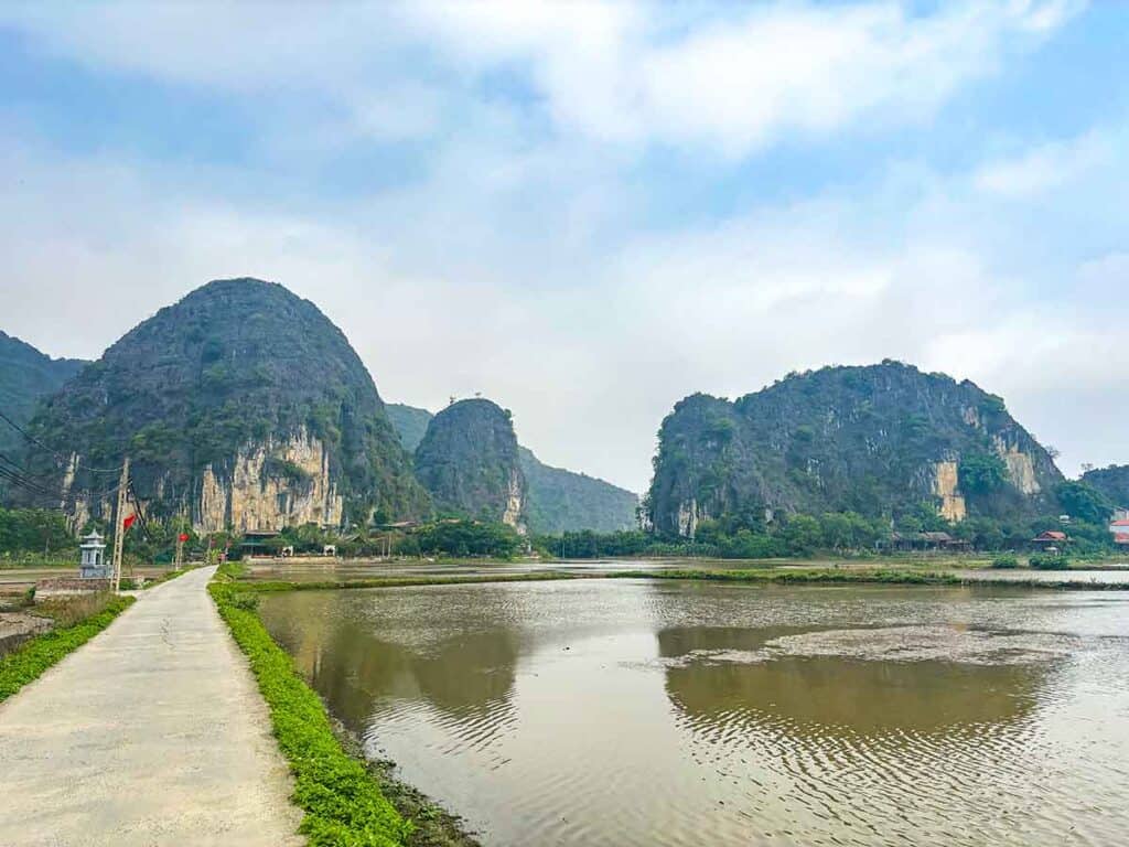 A quiet country road runs beside a tranquil river with towering limestone karsts on either side, in the serene Vietnamese landscape of Tam Coc.