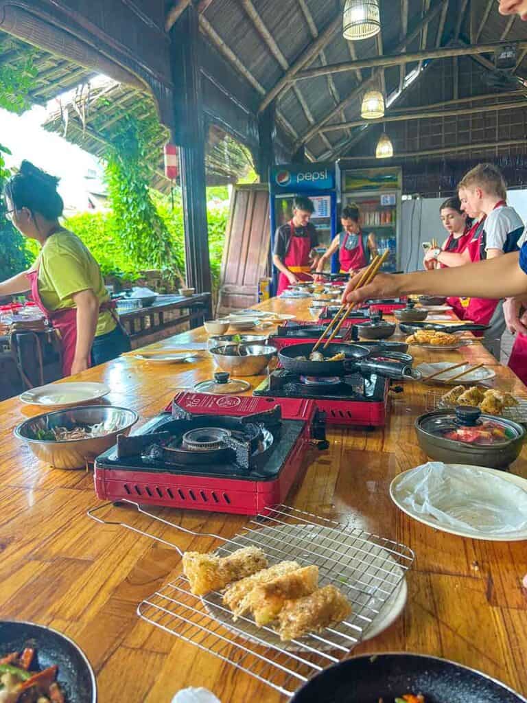 A vibrant Vietnamese cooking class in action with tourists and a local instructor around wooden tables, preparing traditional dishes on red portable burners.